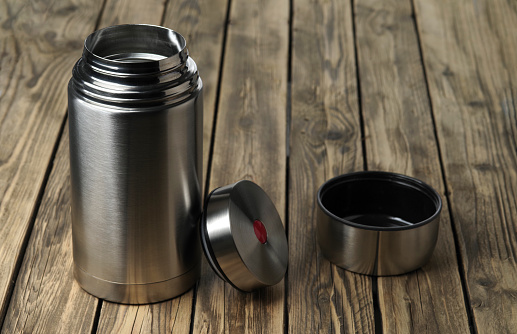 A Stainless-steel vacuum flask with lid removed stands on a wooden table. Selective focus.
