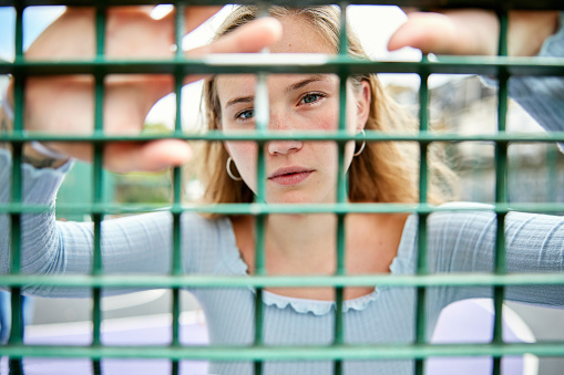 Personal perspective of early 20s blond adult standing with hands on other side of sports fence, focus on face looking at camera with serious expression.
