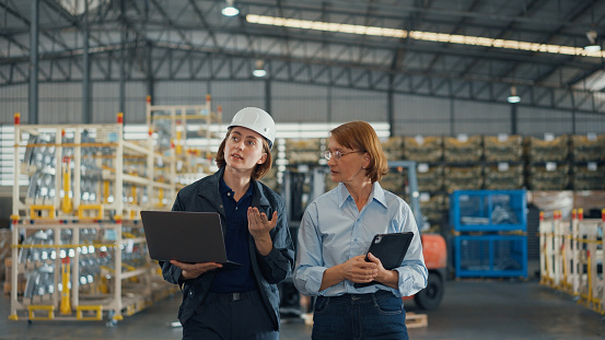 Young caucasian woman worker and mature female manager walking checks stock inventory with tablet and laptop and discuss talk together in retail warehouse. Logistic industry business concept.