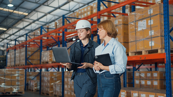 Young caucasian woman worker and mature female manager walking checks stock inventory with tablet and laptop and discuss talk together in retail warehouse. Logistic industry business concept.