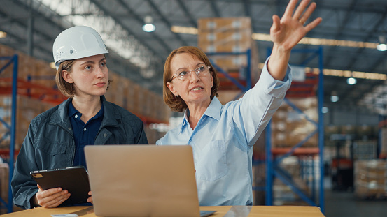 Young caucasian woman worker and mature female manager checks stock inventory with tablet and laptop and discuss talk together in retail warehouse. Logistic industry business concept.