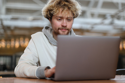 A bearded blond man works behind a laptop indoors.