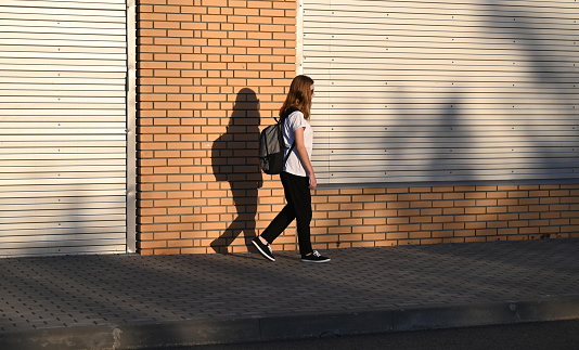 A teenage girl in black trousers and a white T-shirt walks down the street with a black backpack on her back