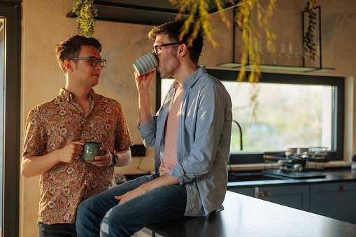 A gay man is sitting on the counter top while his partner is standing next to him and drinking coffee together.