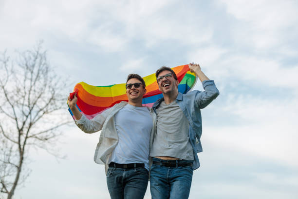 An affectionate homosexual couple is outdoors walking and hugging, holding a rainbow flag above their heads and smiling.