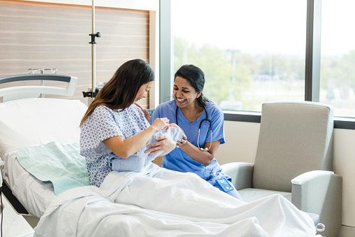 The female nurse sits close to the new mother to reassure her while she tries to breastfeed.