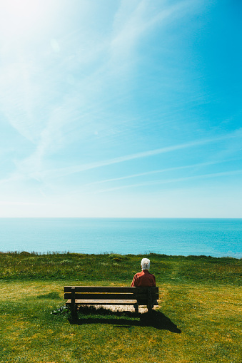 One senior man in his 70s sitting alone on a bench in nature, looking at the view of the sea and Seven Sisters cliffs on the south coast of England.