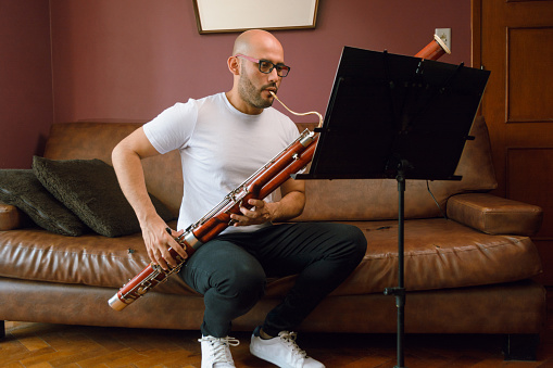 young latino musician man, bald with glasses and white t-shirt, at home studying classical music playing the bassoon sitting on the sofa in the living room, reading sheet music