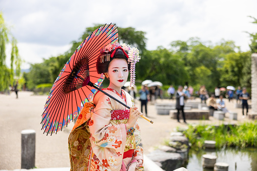 Portrait of Japanese Maiko (Geisha in training) standing in public park in Gion, Kyoto, holding traditional Japanese paper umbrella for sunshade