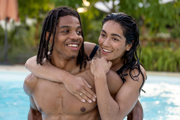 A close up front view of a couple who are enjoying a getaway to Toulouse in the south of France. They are frolicking in the pool and the young woman has her arms wrapped around her partner as they swim in the pool
