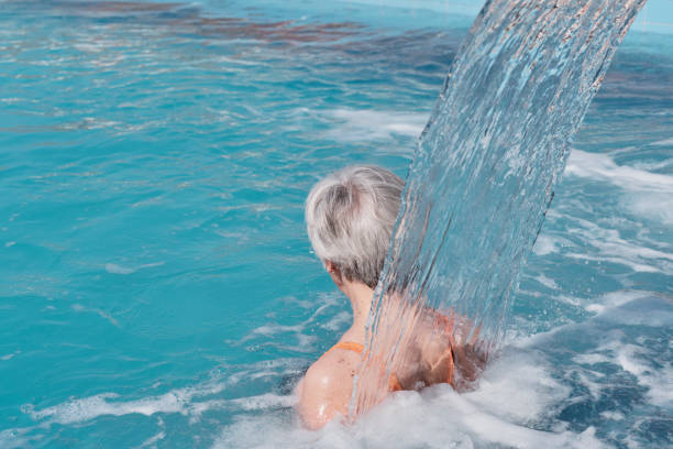 donna anziana con i capelli grigi sotto il flusso dell'acqua che cade dall'alto. veduta posteriore. - physical therapy hydrotherapy swimming healthcare and medicine foto e immagini stock