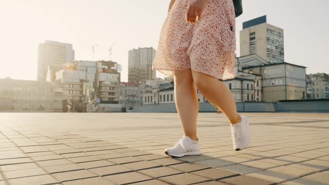 Women's legs in white sneakers slowly walk along the city street.