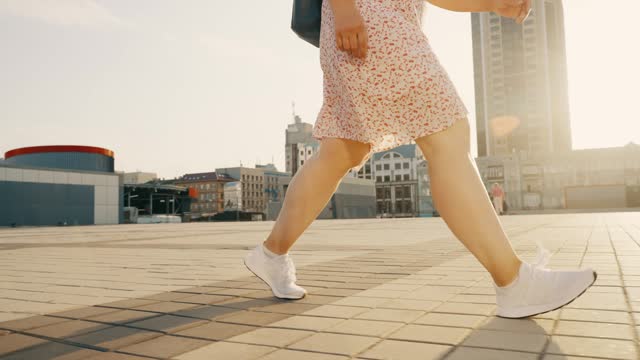 Women's legs in white sneakers go along the sidewalk of a city street.
