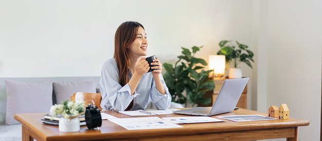 Female freelance is holding coffee to drinking and looking outside to take a break while working on the table in living room at home office.
