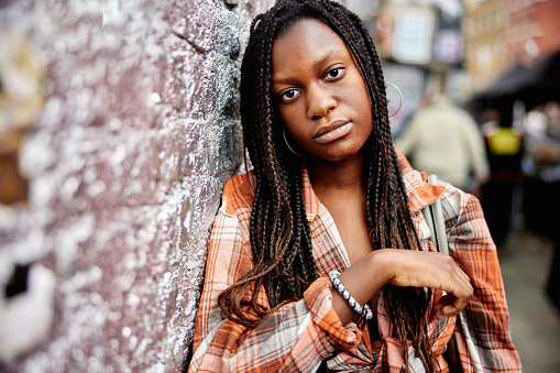 Waist-up portrait of Gen Z adult with long locs wearing plaid blouse and looking at camera with serious expression.