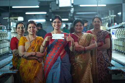 Portrait of confident smiling Indian female manager showing paycheck while standing with coworkers in textile factory and representing women empowerment