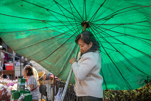 Morning Market, Luang Prabang, Laos - March 15th 2023:  Woman moving a large parasol to create shadow at the morning market which is the main food distribution site in the former capital of Laos