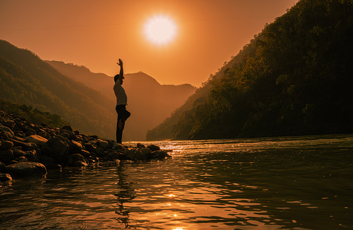 Side view of young man meditating in tree pose while standing on one leg at beautiful lakeshore against mountains and bright sun