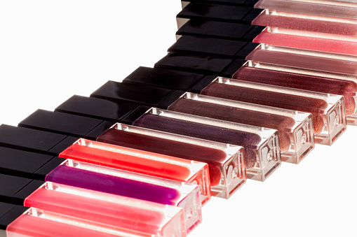 A close-up photograph featuring various shades of lip glosses in transparent containers with black caps. The colors range from pinks, reds to browns, and the containers are arranged in a wave pattern from top-right to bottom-left against a white, backlit background.