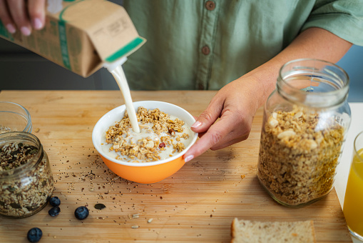 High angle view of woman's hands pouring milk from a carton package into a bowl full of healthy breakfast cereal