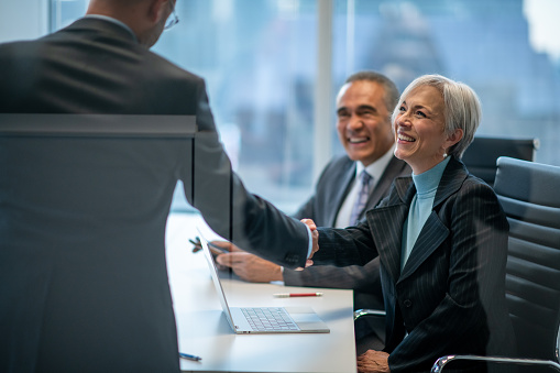 A small group of three business professionals sit together at a boardroom table as they shake hands to solidify a business deal.  They are each dressed professionally and smiling as they happily agree together on the terms.