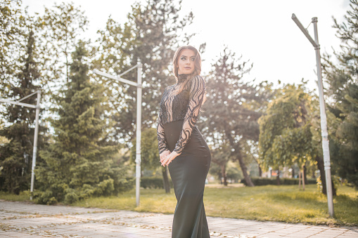 Beautiful modern woman in black long dress with bare back standing outdoors and posing for camera