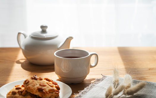 A cup of tea with homemade cookies and a white teapot on a wooden table against the background of a window with sunny morning light and shadow. Early breakfast concept. Free space for text.