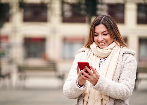 Cheerful young adult woman walking on city street in cold weather and typing a message on her mobile phone - Millennial girl sharing a story on social media platform