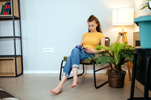 Young woman using mobile phone while sitting in cozy armchair, taking a break and enjoying coffee