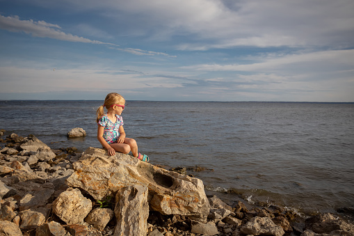 A little girl sits on a stone and looks at the sea. The concept of summer vacation, holidays and vacation at sea. copy space
