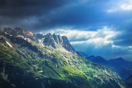 In this photograph, the majestic Mont-Blanc Massif in France is bathed in  dramatic light. The scene unfolds against a backdrop of an ethereal sky, painted in shades of fiery blue and dark grey hues, as the  sun casts its rays through the heavy clouds over the rugged mountain range.
As the light dances upon the landscape, it reveals intricate details and textures. The rugged terrain, marked by deep crevices and jagged edges, tells the story of the immense forces that have shaped these mountains over millions of years.  This landscape photo is a testament to the raw beauty and untamed wilderness of the French Alps, leaving viewers with a deep sense of wonder and an appreciation for the awe-inspiring majesty of nature.