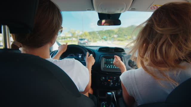 Two young women travel in the car. Cheerful women sit in the modern car listen to a music and dance while travelling on a highway