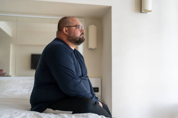 Sad fat lonely man sitting on bed in hotel and looking out window, thinking about life problems Sad fat lonely man sitting on bed in hotel and looking out window, thinking about life problems, no strength change, side view overweight depressed man no friends and mental disorders overweight man stock pictures, royalty-free photos & images