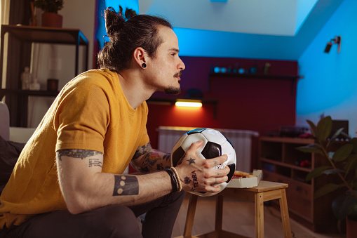 Profile view of a young Cuban man sitting on the couch in the living room and holding a sports ball while watching a football game