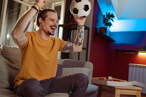 Portrait of a cheerful Latin American man sitting on the couch in the living room and cheering. He is celebrating a goal that his favourite club achieved.