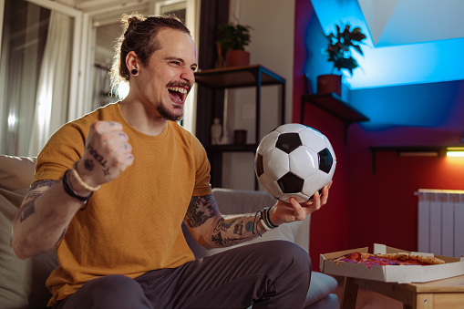 An excited young Latin American man is watching a soccer game at home. He is holding a soccer ball and cheering.