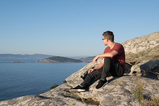 Young man sitting on the rocks of a mountain looking at the ocean