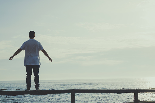 Man with outstretched arms standing on a railing looking at the ocean