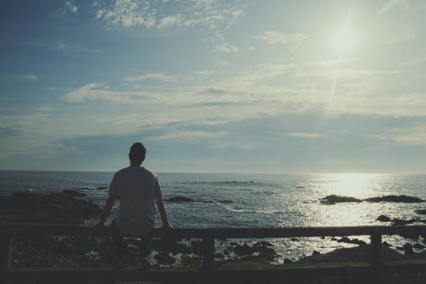 istock Rear view of a man sitting on a railing looking the ocean at sunset 1494928186