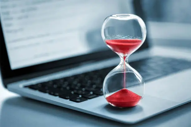 Hourglass with red sand stands on the laptop computer - time management concept