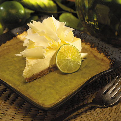 A stylized studio photograph of a slice of homemade Key Lime Pie garnished with white chocolate curls and a slice of lime on a green plate. The plate is resting on a woven mat with a dessert fork in the foreground and limes in the background.