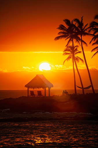 Photo of sunset on a Hawaiian beach with silhouettes of a beach hut, chairs, palm trees and young couple sitting close to one another