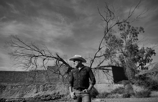 Monochrome of adult man in cowboy hat desert against tree and abandoned building