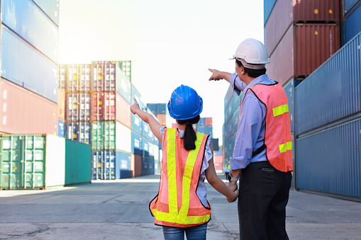 Rear view asian foreman father and his daughter holding hands while teaching little girl. Engineer dad with safety hat point something at container with kid at industrial container cargo freight ship