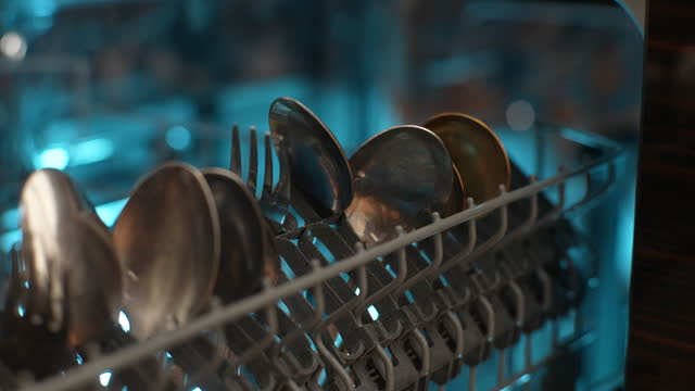 Extreme close-up of unrecognizable housewife loading backlit dishwasher machine with dirty stainless flatware. Concept using modern house kitchen appliance