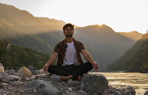 Young handsome man with eyes closed meditating while sitting on rocks at lakeshore against majestic mountains and clear sky during vacation