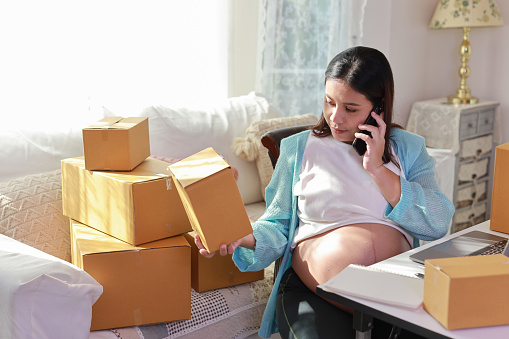 Attractive happy asian pregnant woman sitting and using mobile smart phone and checking order for customer with tablet. Expectant mother preparing and working for baby birth during pregnancy concept.