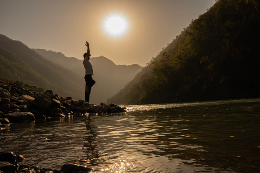 Side view of young man meditating in tree pose while standing on one leg at beautiful lakeshore against mountains and bright sun