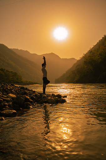 Side view of young man meditating in tree pose while standing on one leg at beautiful lakeshore against mountains and bright sun