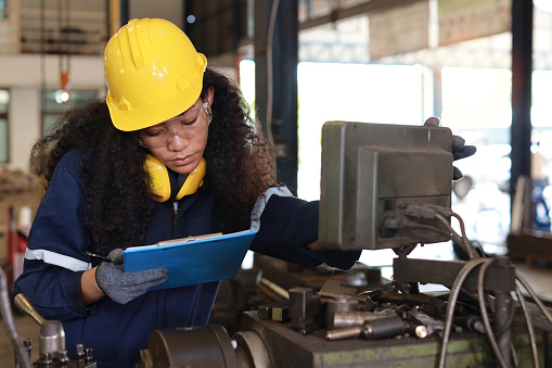 Technician engineer or worker woman in protective uniform and safety hat maintenance operation or check lathe metal machine with clipboard at heavy industry manufacturing factory. Metalworking concept